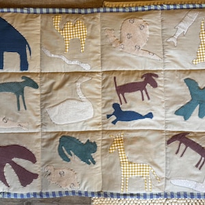 Könnte beinhalten: Eine beige Patchworkdecke mit Applikationen von Tieren in Blau, Grün, Lila und Gelb. Zu den Tieren gehören ein Elefant, ein Kamel, eine Schnecke, ein Fisch, ein Hund, ein Schwan, eine Robbe, ein Vogel, eine Katze, eine Giraffe und ein Fuchs. Die Decke ist mit einem blau-weiß karierten Muster eingefasst.