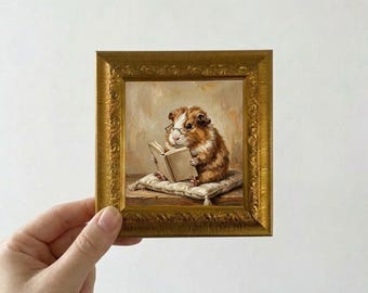 May include: A small, framed painting depicts a guinea pig wearing glasses and reading a book while resting on a pillow. The painting has a gold frame with ornate detailing. The guinea pig is brown and white.