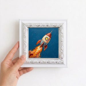 Könnte beinhalten: Ein Ölgemälde einer Rakete, die vor einem tiefblauen Hintergrund aufsteigt, mit einem metallischen Körper, einem gelben Bullauge und leuchtend orangefarbenen sowie roten Flammen, eingefasst in einen verzierten weißen Rahmen.