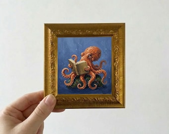May include: A small, square, framed painting featuring an orange octopus reading a book. The octopus is set against a blue background, and the frame is gold. The artwork is held by a hand.