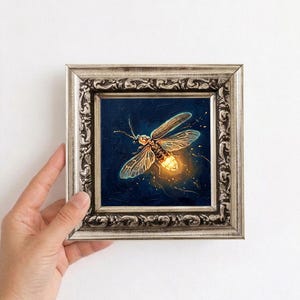 Puede incluir: Pintura enmarcada de una luci&eacute;rnaga con abdomen brillante y alas transl&uacute;cidas. La obra de arte est&aacute; sobre un fondo azul oscuro, y el marco es plateado con detalles ornamentados. La pintura se sostiene sobre un fondo blanco.