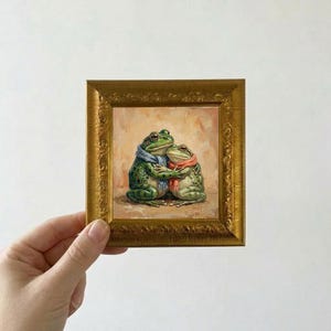 Peut inclure: Peinture encadrée représentant deux grenouilles vertes s'embrassant. L'une porte une écharpe bleue, l'autre une écharpe rouge. L'œuvre est placée sur un fond chaud et neutre, dans un cadre doré orné.