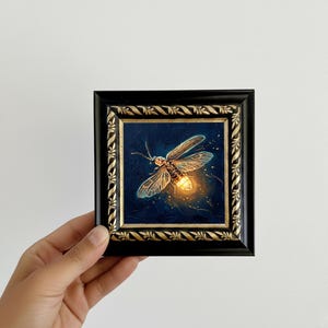 Puede incluir: Pintura enmarcada de una luci&eacute;rnaga sobre un fondo azul oscuro. La luci&eacute;rnaga tiene alas transl&uacute;cidas y un abdomen brillante. El marco es negro con detalles dorados. La obra de arte est&aacute; sujeta por una mano.