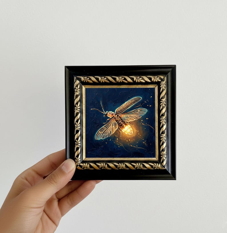Puede incluir: Pintura enmarcada de una luci&eacute;rnaga sobre un fondo azul oscuro. La luci&eacute;rnaga tiene alas transl&uacute;cidas y un abdomen brillante. El marco es negro con detalles dorados. La obra de arte est&aacute; sujeta por una mano.