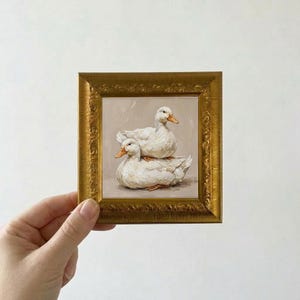 May include: A small square oil painting featuring two white ducks with orange beaks, one resting on top of the other. The artwork is enclosed in an ornate, metallic gold-colored frame with a textured, embossed floral pattern along the border.
