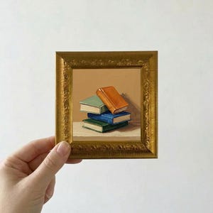 以下が含まれることがあります： 金色の額縁に入った小さな絵画。ミニチュアの本が積み重ねられています。本は緑、青、オレンジ色です。絵画は白い背景を背景に手で持たれています。