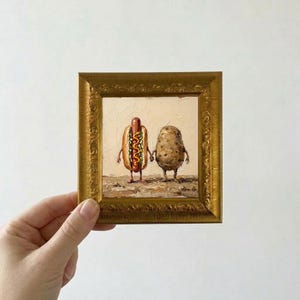 May include: An oil painting in a decorative gold frame depicting a hot dog with mustard and a potato standing side by side on a textured ground. The hot dog and potato have thin arms and legs, and they are holding hands against a cream background.