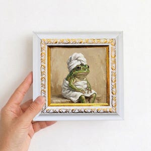 May include: A framed painting depicts a green frog wrapped in a white towel and wearing a towel on its head. The painting is in a white frame with gold accents. The frog is sitting on a bench.