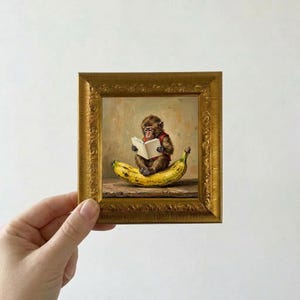 Op de afbeelding: Een klein ingelijst olieverfschilderij met een bruine aap in een rood vestje, zittend op een grote gele banaan terwijl hij een open wit boek leest. Het kunstwerk zit in een sierlijke, goudkleurige lijst met gedetailleerd snijwerk.