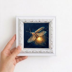 Puede incluir: Pintura enmarcada de una luci&eacute;rnaga con abdomen brillante y alas transl&uacute;cidas. La obra de arte est&aacute; sobre un fondo azul oscuro con un marco blanco ornamentado. La pintura est&aacute; sostenida por una mano.