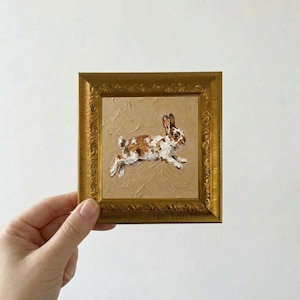 Puede incluir: Pequeña pintura al óleo enmarcada de un conejo corriendo. El conejo es blanco con manchas marrones sobre un fondo beige. El marco es dorado y ornamentado.