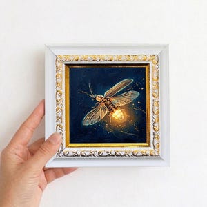 Puede incluir: Pintura enmarcada de una luci&eacute;rnaga sobre un fondo azul oscuro. La luci&eacute;rnaga se representa con alas transl&uacute;cidas y un abdomen brillante. El marco blanco con detalles dorados a&ntilde;ade un toque de elegancia a la obra.