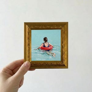 May include: A framed painting depicts a person in a blue swimsuit with a red inflatable ring in a body of water. The painting is in a gold-colored ornate frame. The water is a light blue color.