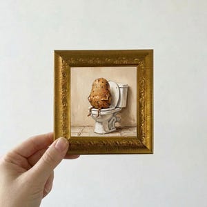 Könnte beinhalten: Ein kleines quadratisches Gemälde in einem verzierten Goldrahmen, das eine braune, texturierte Kartoffelfigur auf einer weißen Porzellan-Toilette zeigt. Das Kunstwerk hat einen neutralen beigen Hintergrund und einen Fliesenboden.