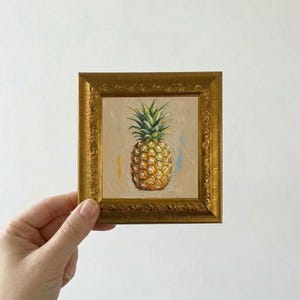 Puede incluir: Pequeña pintura cuadrada de una piña en un marco dorado ornamentado. La piña es amarilla y texturizada, con hojas verdes. El fondo es beige neutro con pinceladas azules y amarillas.
