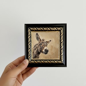 May include: A square framed oil painting featuring a detailed portrait of a brown donkey against a neutral beige background. The frame is black with ornate gold-coloured carved detailing along the inner border, creating a distinct contrast.
