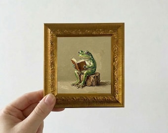 May include: A framed painting depicts a green frog reading a book while seated on a tree stump. The artwork is set within a gold-colored, ornate frame. The frog is the focal point, with detailed rendering of its features and the book.