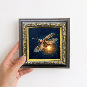 Puede incluir: Pintura enmarcada de una luci&eacute;rnaga sobre un fondo azul oscuro. La luci&eacute;rnaga est&aacute; representada con luz amarilla y naranja, con alas y cuerpo detallados. El marco es oscuro con detalles dorados.