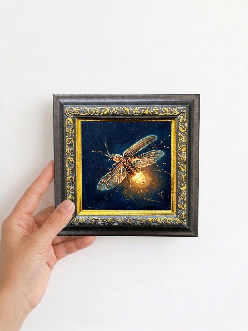 Puede incluir: Pintura enmarcada de una luci&eacute;rnaga sobre un fondo azul oscuro. La luci&eacute;rnaga est&aacute; representada con luz amarilla y naranja, con alas y cuerpo detallados. El marco es oscuro con detalles dorados.
