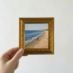 Könnte beinhalten: Ein kleines, quadratisches, gerahmtes Gemälde, das eine Strandszene mit zwei Personen zeigt, die am Ufer entlang gehen. Das Gemälde hat einen goldenen, verzierten Rahmen. Das Meer ist blau und der Sand ist hellbraun. Der Himmel ist hellblau.