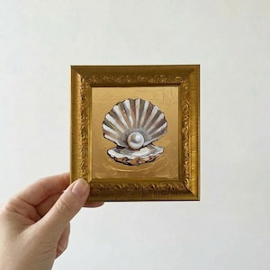May include: A small, square painting in a gold frame depicts a seashell with a pearl. The shell is white and brown, set against a gold background. The frame has an ornate, textured design.