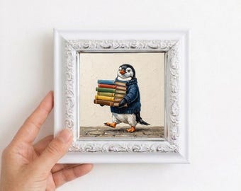 May include: A framed square print featuring an illustration of a penguin wearing a dark blue knitted sweater and carrying a stack of five colorful books. The artwork is encased in a white ornate frame with a detailed, textured decorative border.