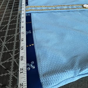 May include: A light blue fabric with a gold chain trim and blue trim with rhinestones. The fabric is measured with a ruler that shows 22 inches.