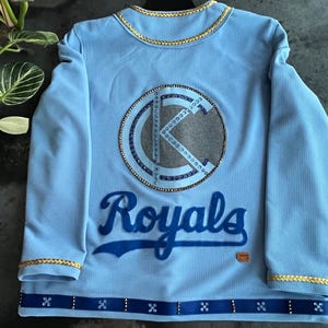 May include: Light blue long-sleeved jersey with a large, sparkly, white and blue "K" logo and the word "Royals" in blue lettering.