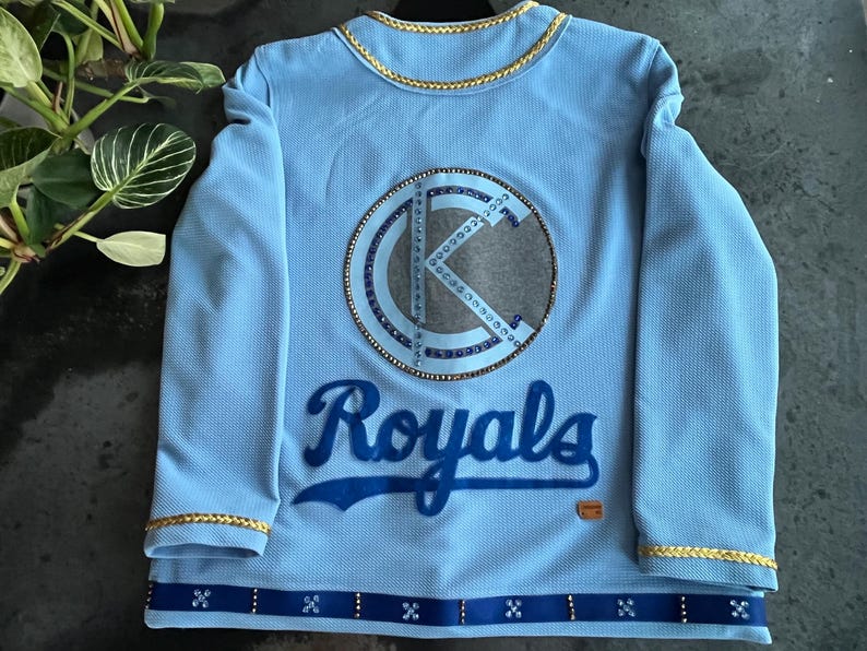 May include: Light blue long-sleeved jersey with a large, sparkly, white and blue "K" logo and the word "Royals" in blue lettering.