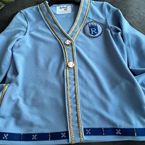 May include: A light blue cardigan with gold trim and a blue and white baseball-themed patch with the letter "R" in a crown. The cardigan has a button closure and a blue ribbon with rhinestones around the bottom.