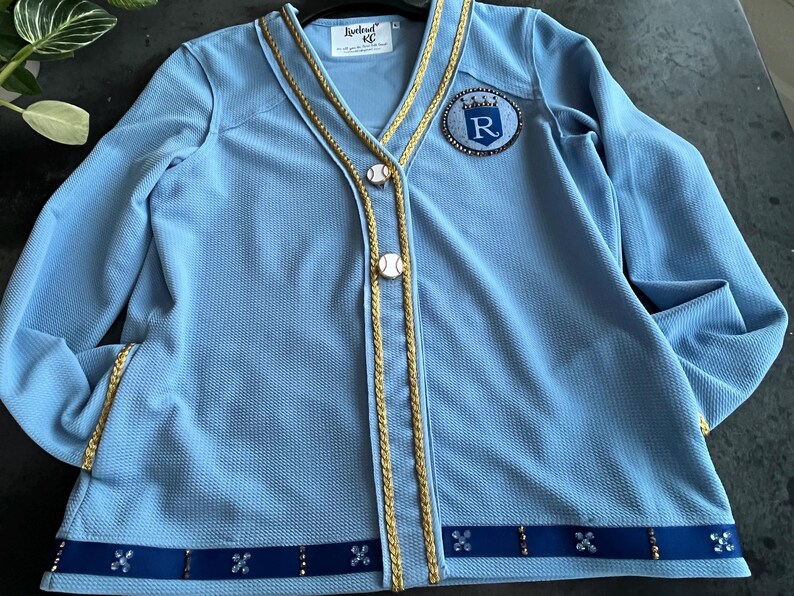 May include: A light blue cardigan with gold trim and a blue and white baseball-themed patch with the letter "R" in a crown. The cardigan has a button closure and a blue ribbon with rhinestones around the bottom.