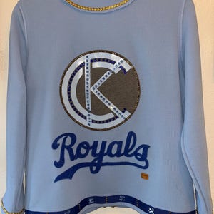 May include: A light blue jacket with a gold chain trim. The back of the jacket features a large, rhinestone-encrusted "K" logo with a brown background. Below the logo is the word "Royals" in blue script.