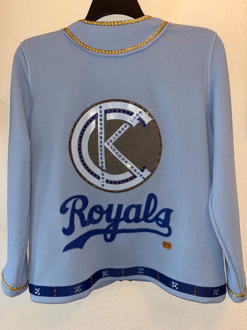 May include: A light blue jacket with a gold chain trim. The back of the jacket features a large, rhinestone-encrusted "K" logo with a brown background. Below the logo is the word "Royals" in blue script.