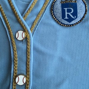 May include: A light blue baseball jersey with gold braid trim and a white baseball button. The jersey has a blue and white patch with a crown and the letter "R" on the right side.