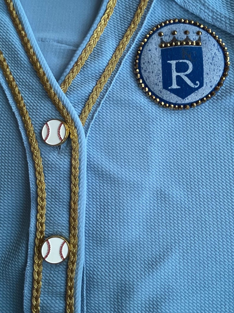 May include: A light blue baseball jersey with gold braid trim and a white baseball button. The jersey has a blue and white patch with a crown and the letter "R" on the right side.