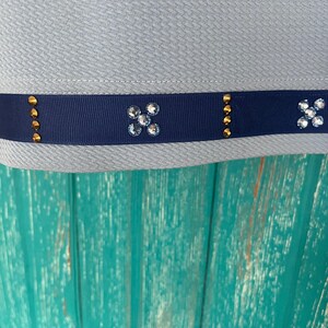 May include: A light blue fabric with a navy blue ribbon trim. The ribbon has gold accents and clear rhinestones in a flower shape.