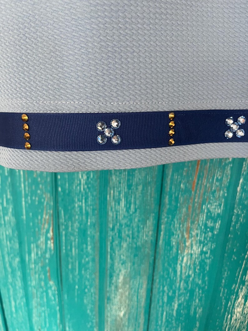 May include: A light blue fabric with a navy blue ribbon trim. The ribbon has gold accents and clear rhinestones in a flower shape.