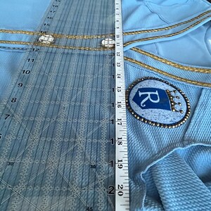 May include: A light blue garment with gold trim and a blue and gold embroidered patch with the letter "R" on it. The garment is being measured with a ruler.