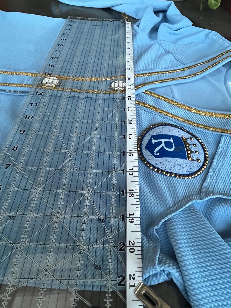 May include: A light blue garment with gold trim and a blue and gold embroidered patch with the letter "R" on it. The garment is being measured with a ruler.