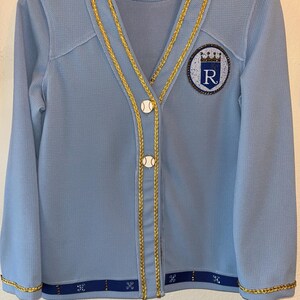 May include: A light blue cardigan with gold trim and a baseball-themed button. The cardigan has a large embroidered patch on the left side featuring a royal blue circle with a crown and the letter "R" in rhinestones. The text "LiveLoud KC" is on a white tag attached to the cardigan.