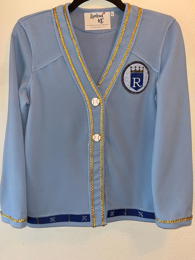 May include: A light blue cardigan with gold trim and a baseball-themed button. The cardigan has a large embroidered patch on the left side featuring a royal blue circle with a crown and the letter "R" in rhinestones. The text "LiveLoud KC" is on a white tag attached to the cardigan.