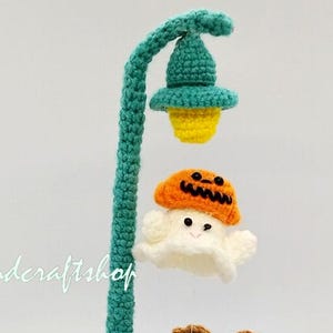 Puede incluir: Un farol de ganchillo hecho a mano con un poste verde azulado y una base rosa. La lámpara presenta una luz amarilla, un fantasma con un sombrero de calabaza y un pequeño búho. El texto "Crochet Pattern" está en la parte superior.