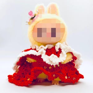 May include: A plush toy wearing a yellow bunny hood with a pink flower crown and a red crocheted cape with white trim. The cape is adorned with gold sequins. The toy has a blurred face.