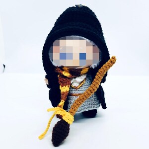 Può includere: Bambola all'uncinetto fatta a mano vestita con un mantello con cappuccio nero, una sciarpa a righe marroni e gialle e un completo grigio. La bambola tiene in mano una scopa marrone e gialla. Il viso della bambola è pixelato.