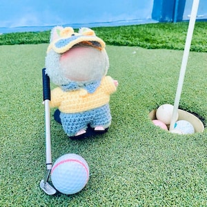 May include: A crocheted doll dressed in a yellow sweater, blue shorts, and a matching hat, holding a miniature golf club. A golf ball with red stripes rests on the green grass, near the hole with other golf balls. The doll has a blank face.