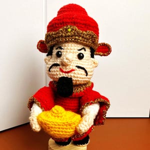 以下が含まれることがあります： 赤いローブと金の冠をかぶった中国の神様の編みぐるみ。金の延べ棒を持っています。編みぐるみは木製の台座の上に立っています。