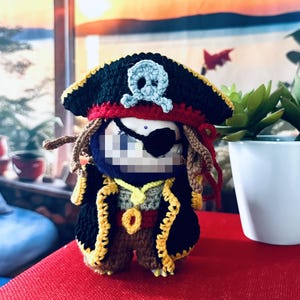 Pode incluir: Boneca pirata de crochê feita à mão. Possui um chapéu preto com uma caveira e ossos cruzados azuis claros, tapa-olho preto e rastas castanhas. A boneca usa um casaco preto com acabamento amarelo e um colar dourado.