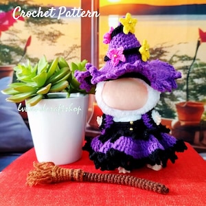 Puede incluir: Una muñeca de ganchillo hecha a mano con un sombrero y vestido de bruja morados, con una pequeña escoba. La muñeca tiene una cara blanca y está decorada con adornos en forma de estrella rosa y amarilla. El texto "Crochet Pattern" es visible.