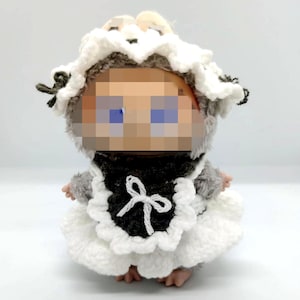 May include: A handmade doll dressed in a white and black crocheted maid outfit. The doll has gray fur and a white bonnet. The outfit includes a white apron with a black bib and a white bow. The doll's face is pixelated.