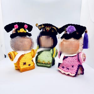 Könnte beinhalten: Drei Plüschfiguren in farbenfrohen, gehäkelten traditionellen Outfits. Die Figuren tragen schwarze Hüte mit Blumen und goldenen Akzenten. Die Outfits sind gelb, grün und rosa, jedes mit einzigartigen Details. Auf weißem Hintergrund.
